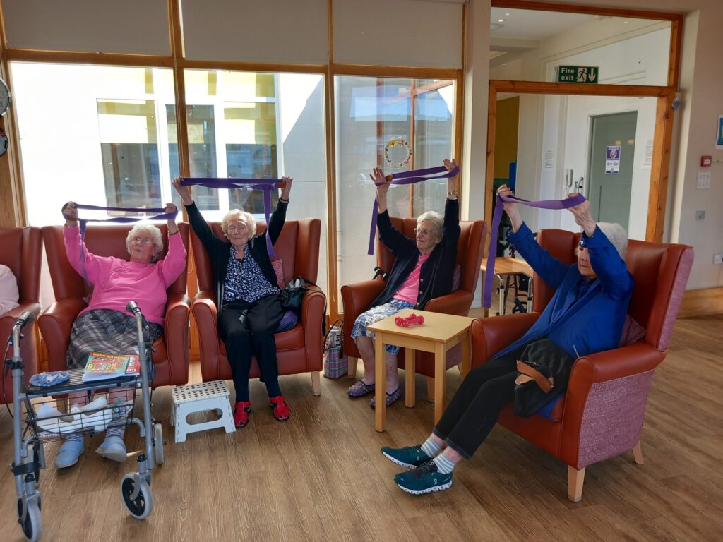Four elderly women enjoy an Active Afternoon, raising exercise bands above their heads in a bright room with large windows.