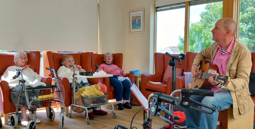 A man plays the guitar and sings songs to three elderly women seated in chairs, two of whom have walking frames and baskets, in a bright room with large windows and plenty of smiles.