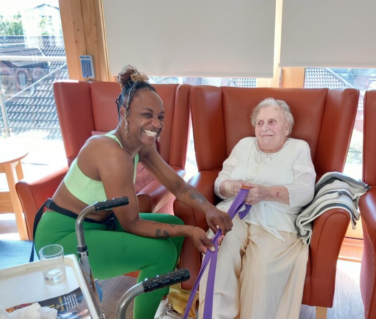 Carla smiles in her workout clothes as she helps an elderly woman with a resistance band exercise—just one of many Active Afternoons enjoyed in the brightly lit room with red chairs.