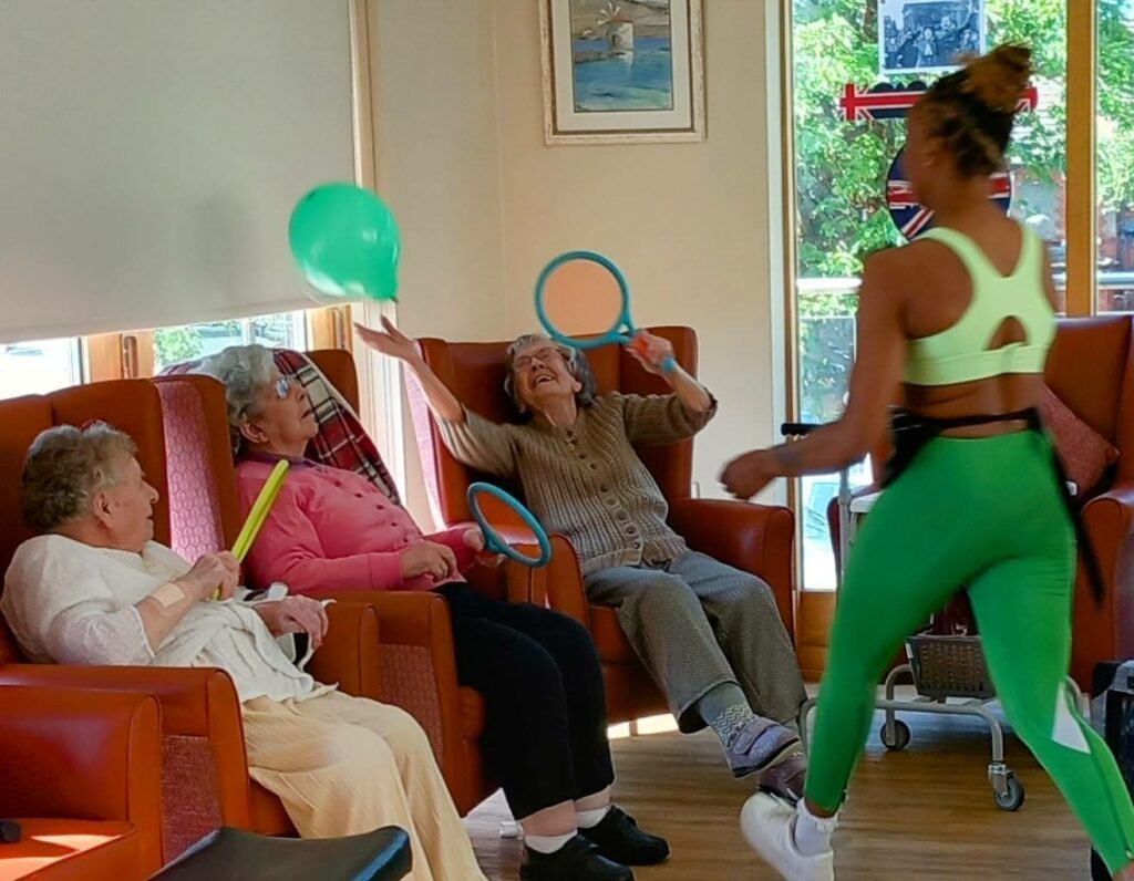 Three elderly women enjoy an Active Afternoons balloon game with rings, led by an instructor in green sportswear, filling the bright room with laughter and smiles.