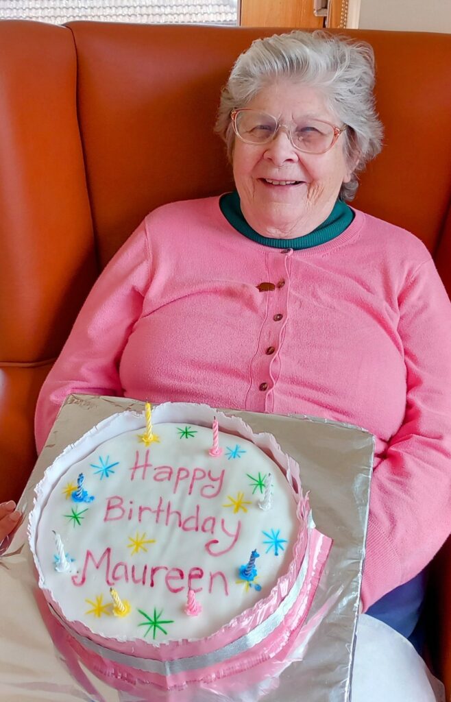 An elderly woman wearing glasses and a pink jumper smiles as she holds a decorated birthday cake with ‘Happy Birthday Maureen’ written on it, ready for songs and celebration.