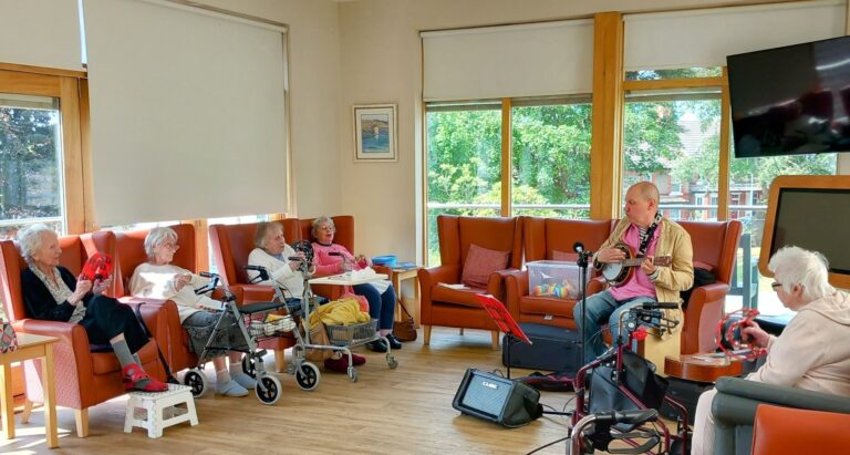 A group of older adults sit in a sunlit room on armchairs, some with walking frames, watching a man play songs on a banjo. Musical instruments and a music stand are visible, and smiles light up their faces.