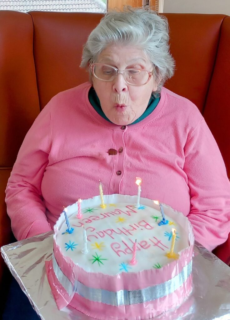 An elderly woman in a pink jumper blows out candles on a decorated birthday cake with Happy Birthday written on it, surrounded by smiles.
