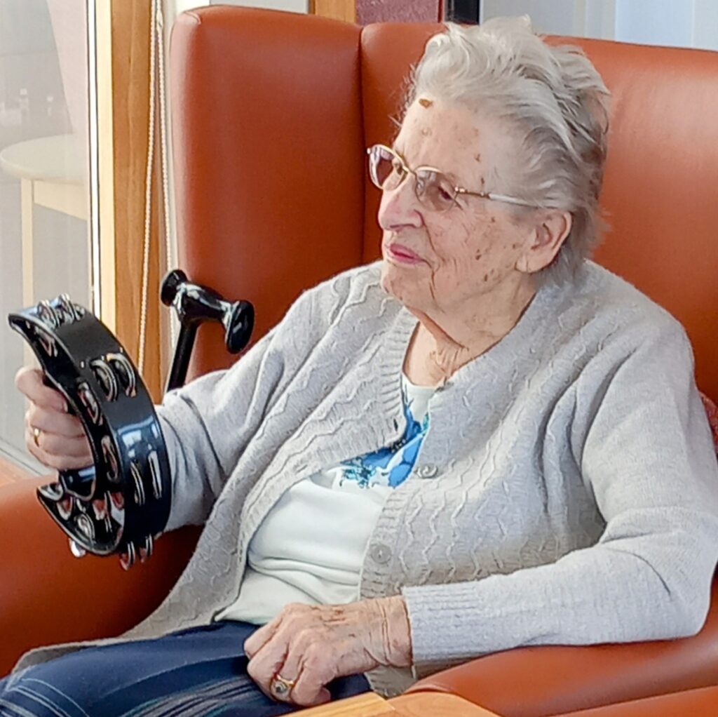 Elderly woman smiles as she sits in an armchair, holding a tambourine in her right hand, wearing glasses, a grey cardigan, and checked trousers.