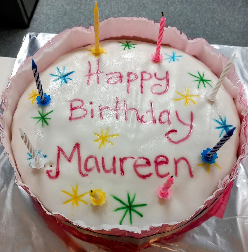 Round birthday cake with white icing, colourful star decorations, six unlit candles, and “Happy Birthday Maureen” written in pink icing—perfect for smiles and songs on her special day.
