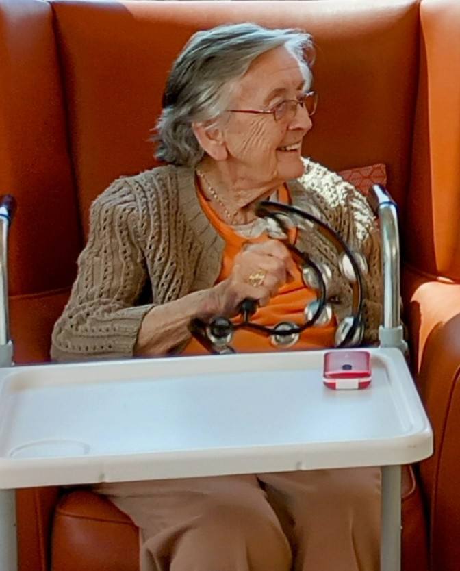 An elderly woman sits in an orange armchair, smiling and holding a tambourine. A white tray with a pink cake is in front of her, adding to the cheerful scene. In British English: An elderly woman sits in an orange armchair, smiling and holding a tambourine. A white tray with a pink cake is placed in front of her, adding to the cheerful atmosphere. (There are no significant changes needed except potentially swapping scene for atmosphere, which is more common in British descriptions. The spelling and terms used are already suitable.).