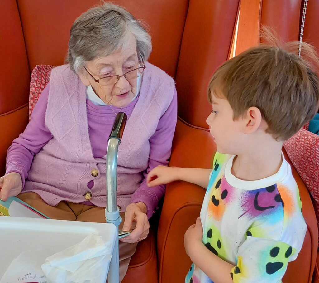 An elderly woman in a lavender jumper sits in a chair, chatting with a young boy from St Agnes Pre-School who stands beside her.