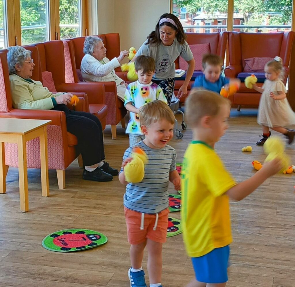 Young children and elderly women from the St Agnes Pre-School community interact and play with yellow toys in a brightly lit room furnished with armchairs and large windows.