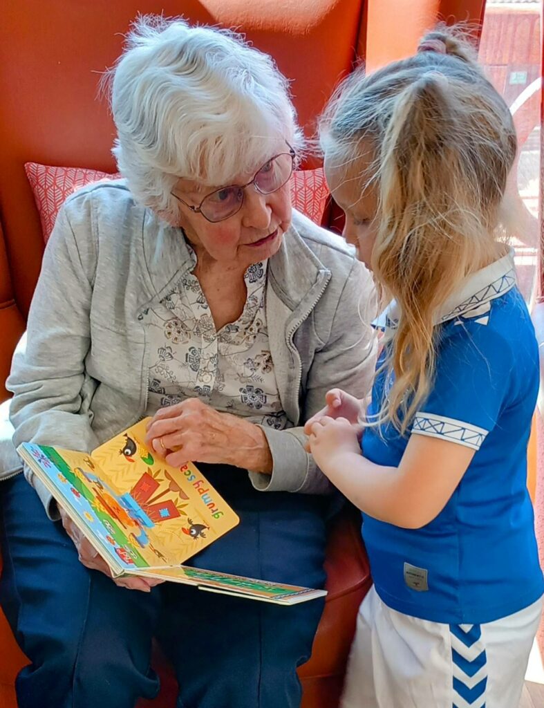 An elderly woman reads a colourful children’s book to a young girl standing beside her in a bright, cosy room at St Agnes Pre-School, creating a warm sense of community.
