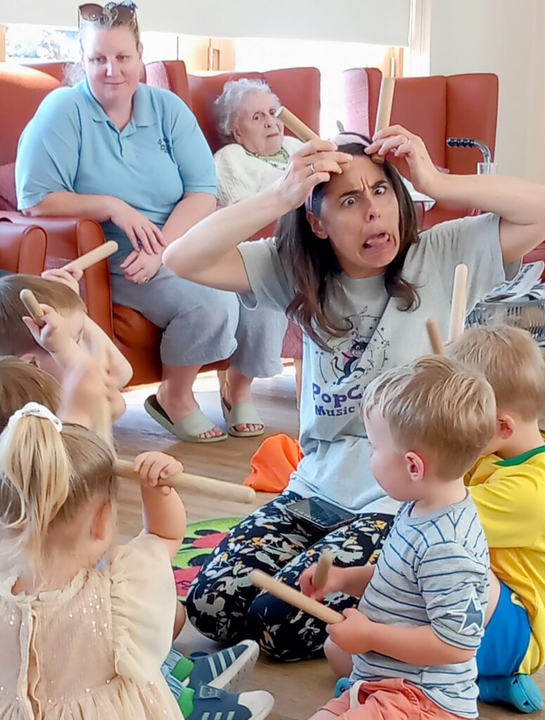 A woman pulls a funny face while sitting on the floor with young children holding sticks at St Agnes Pre-School, as two older women from the community watch from chairs in the background.
