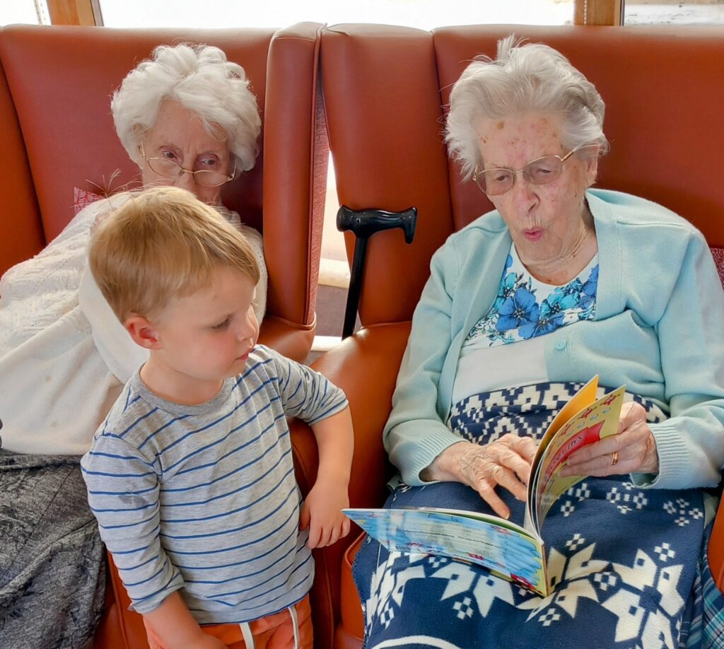 Two elderly women are seated in armchairs; one is reading a book to a young boy from the St Agnes Pre-School community, while a walking stick rests between them.