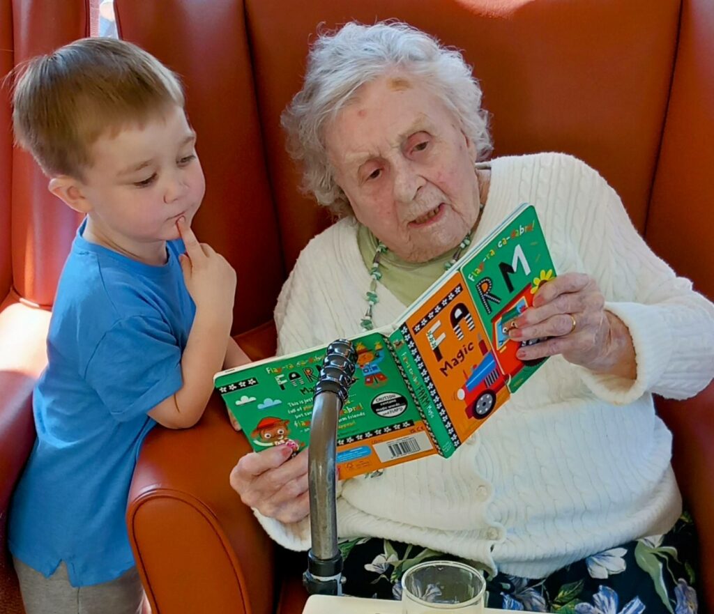 An elderly woman sits in a chair reading a children’s book to a young boy at St Agnes Pre-School, both engaged and demonstrating the warmth of community learning together.