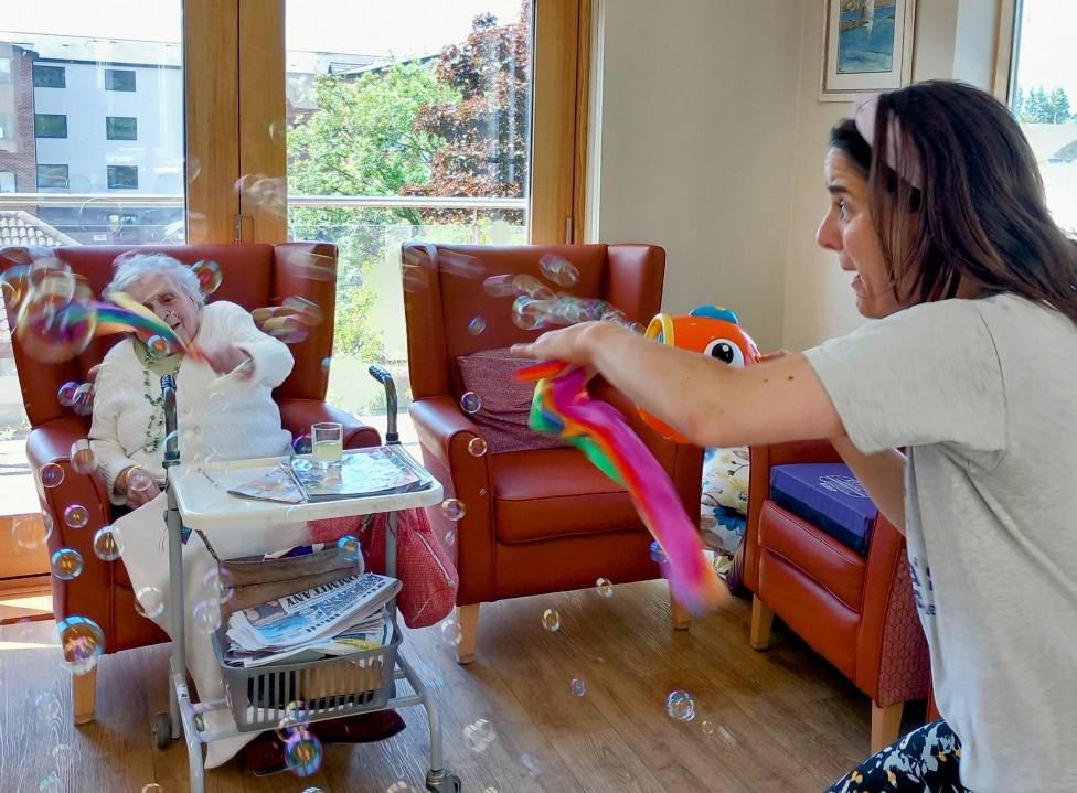 A young woman from St Agnes Pre-School waves colourful ribbons to create bubbles, entertaining an elderly woman in a wheelchair inside a bright community room with large windows and red chairs.