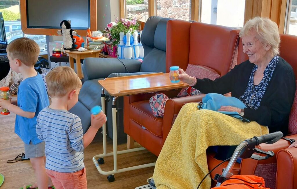 Two young children from St Agnes Pre-School play with toy shakers in front of an elderly woman, sharing smiles and joy in a brightly lit community room.