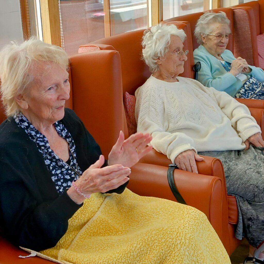 Three elderly women sit in armchairs by a window, wrapped in blankets. One claps her hands while the others rest theirs on their laps, sharing a moment of togetherness reminiscent of St Agnes Pre-School gatherings.
