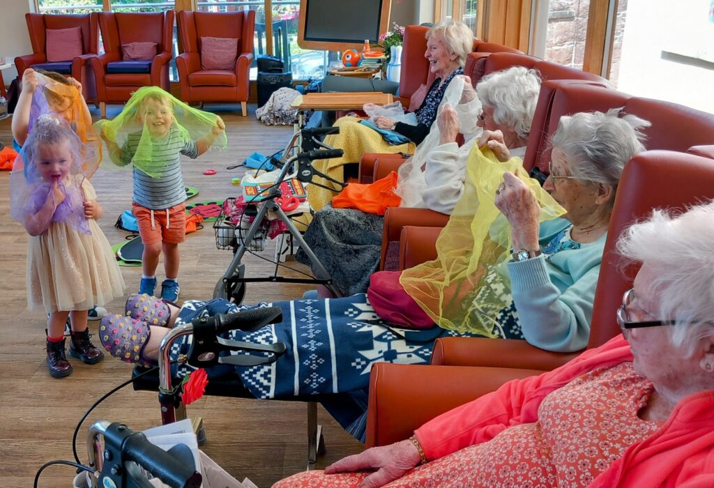 Children and elderly women from the community play together indoors at St Agnes Pre-School, smiling and holding colourful scarves amid chairs, windows, and scattered items on the floor.