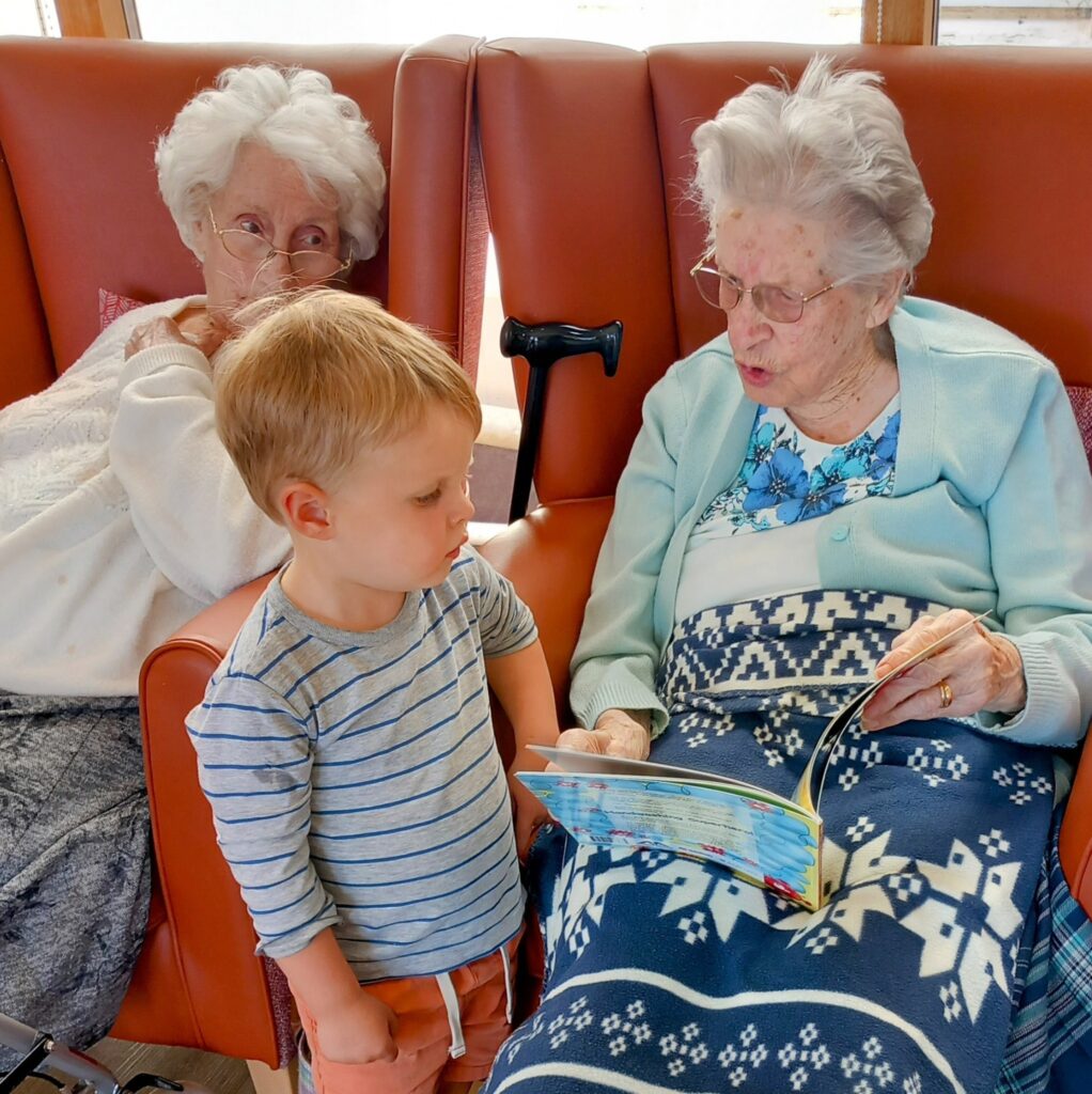 An elderly woman from the St Agnes Pre-School community reads a book to a young boy, while another elderly woman sits nearby in an armchair, watching them.