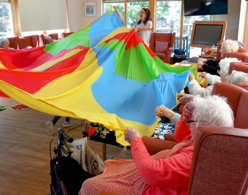 In a cosy community lounge, a group of elderly people hold the edges of a large, colourful parachute, guided by a woman from St Agnes Pre-School standing in the background.