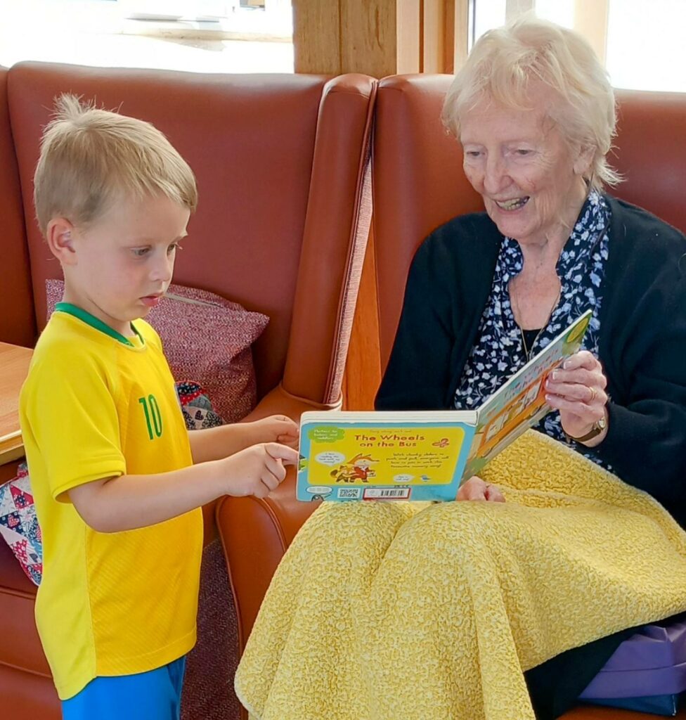 A young boy in a yellow shirt and an elderly woman sit together, reading a children’s book at St Agnes Pre-School, sharing a special community moment as the boy points to the page.