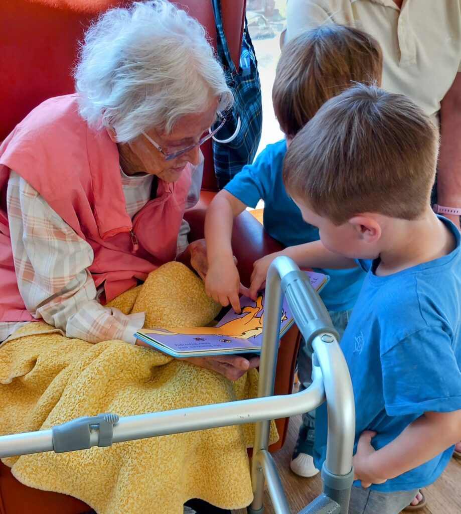 An elderly woman sits with a blanket and a walking frame, reading a picture book to two young boys from the St Agnes Pre-School community, who are eagerly pointing at the pages.