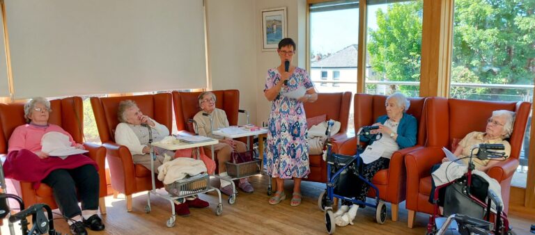 On a Thoughtful Afternoon, the Reader stands and recites poetry from a sheet of paper while five elderly women sit in armchairs—some holding papers or using walking frames—in a brightly lit room.