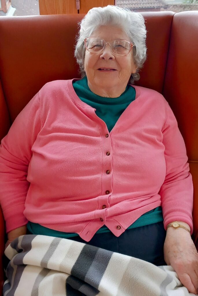 Elderly woman with white hair and glasses, wearing a pink cardigan and green top, sits in a chair with a striped blanket on her lap, gently knitting as she enjoys a quiet afternoon.