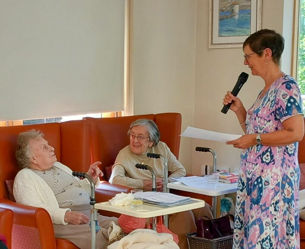 Three elderly women are in a room; two with walking frames chat, while the Reader stands holding a microphone and papers, sharing poetry that stirs cherished memories among her attentive friends.