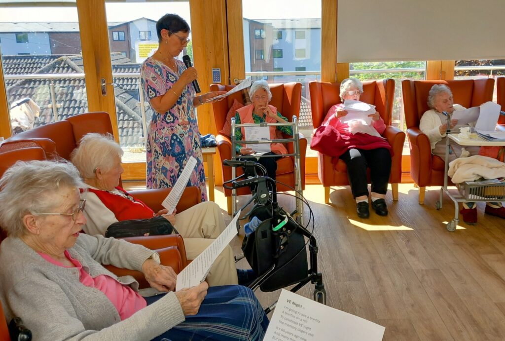 A group of elderly women sit in a sunlit room reading pages of poetry, while the reader stands at the front holding a sheet of paper and a microphone.