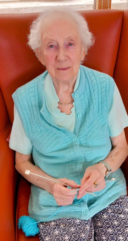 Elderly woman with short white hair sits on a red chair, wearing a light blue waistcoat and patterned trousers, knitting with needles and wool.