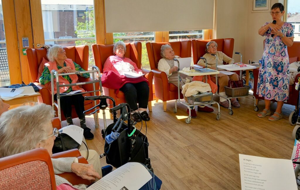 A group of elderly women sit in armchairs in a sunlit room, listening to the reader speaking while holding a microphone; pages of poetry rest on their laps and tables.