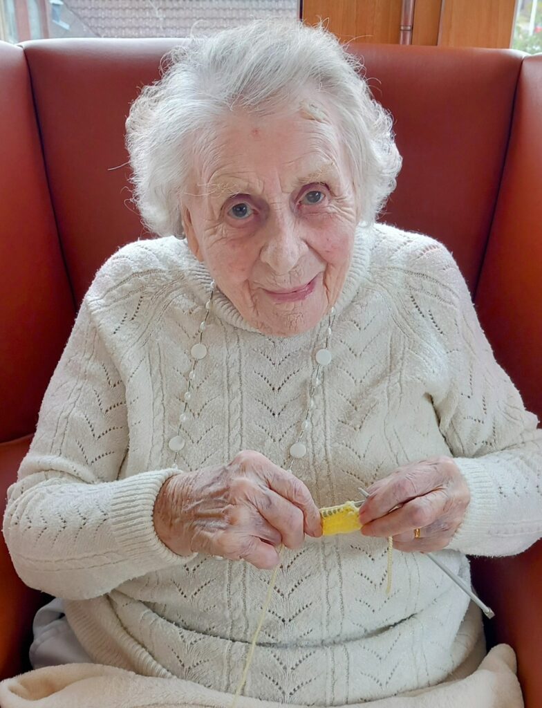 An elderly woman with white hair sits in an orange chair, wearing a cream jumper, knitting with yellow wool. She looks at the camera—focused and enjoying the gentle click of her needles.