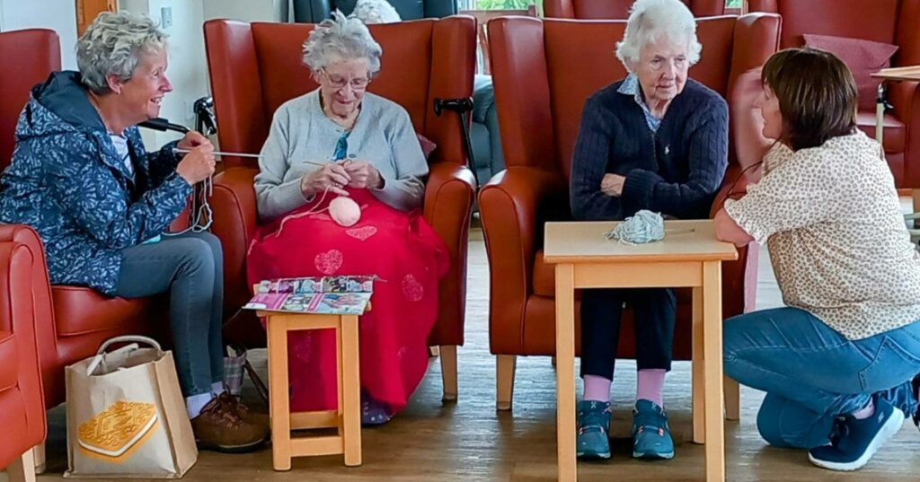 Four women in a bright lounge, three seated on armchairs with knitting supplies and one kneeling beside them, all happily engaged in conversation and nattering.