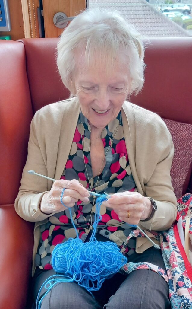 An elderly woman sits on a red sofa, smiling as she knits with blue wool and knitting needles, enjoying the peaceful clatter of her craft.