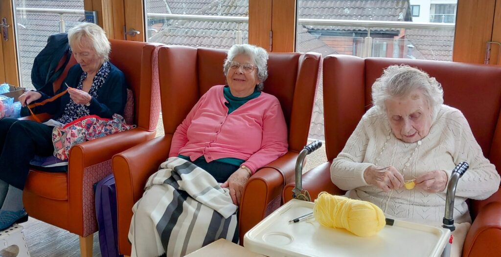 Three elderly women sit in armchairs by a window; one is knitting, one smiles at the camera, and one crochets with yellow wool.