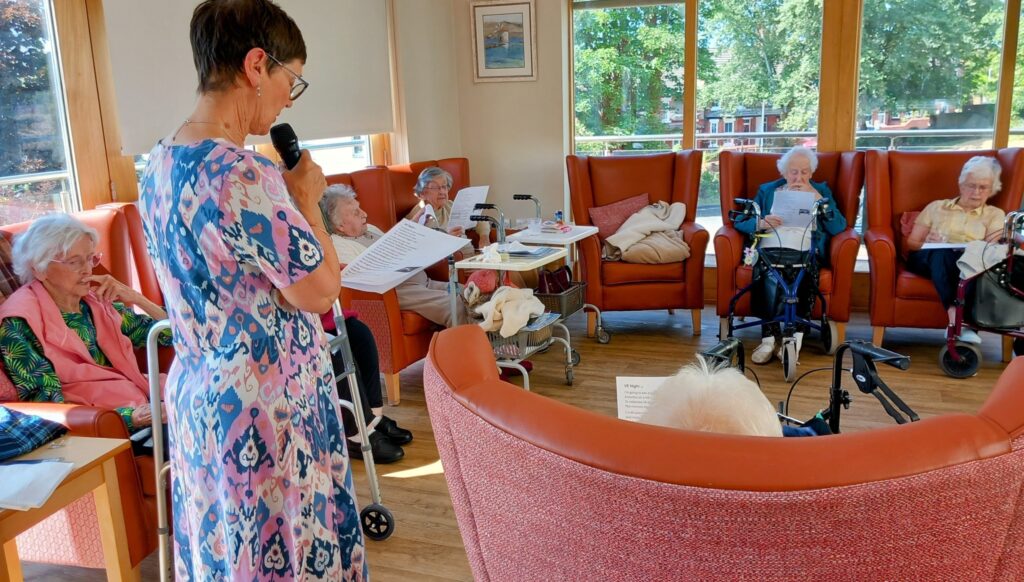 A woman stands and speaks into a microphone, sharing poetry, while elderly people sit in armchairs holding sheets of paper in a bright communal room with large windows.