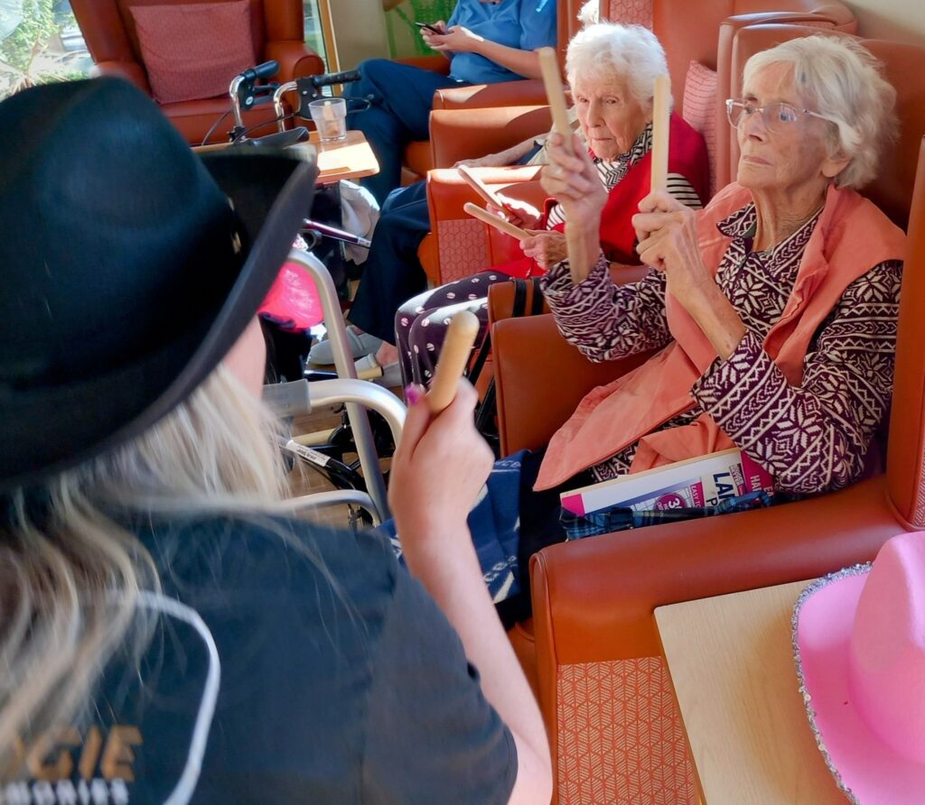 Two elderly women seated in chairs hold wooden rhythm sticks, facing a person in a black hat, during an indoor Yee-Haw group activity.