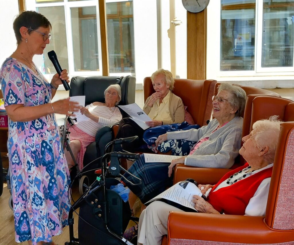 A woman stands holding a microphone, sharing poetry with four elderly women seated in armchairs, smiling and holding papers in a brightly lit room. Heres the rewritten version in British English: A woman stands holding a microphone, reciting poetry to four elderly women seated in armchairs, smiling and holding sheets of paper in a brightly lit room.