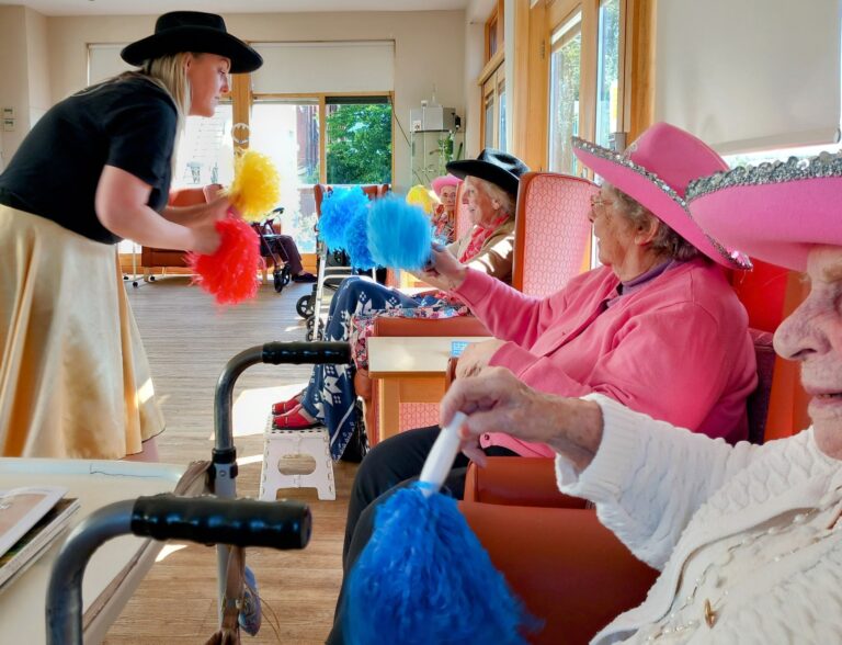 A group of elderly women in hats sit in chairs, holding colourful pom-poms, while a woman stands in front of them, leading a lively Yee-Haw themed activity in a sunlit room.