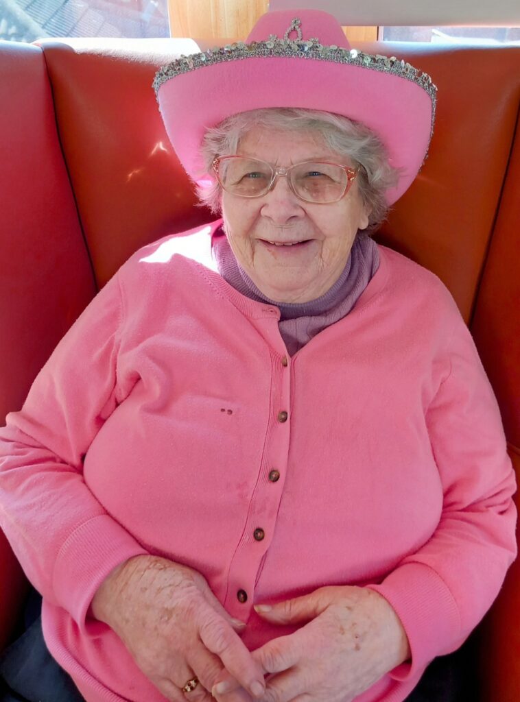 Elderly woman with glasses smiling, wearing a pink jumper and a pink hat adorned with a tiara, seated on a red cushioned bench, exuding real Yee-Haw charm.