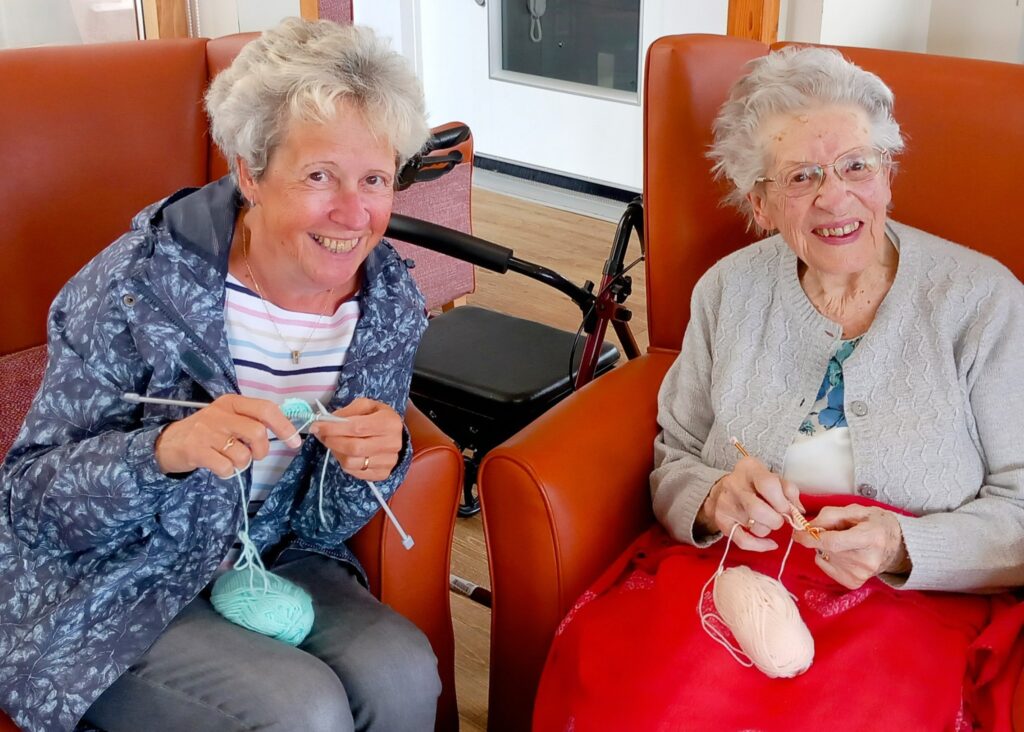 Two older women sit in armchairs, smiling and nattering as they knit with pastel wool. A walking frame is visible in the background.