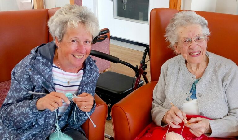 Two older women sit in armchairs, smiling and nattering as they knit with pastel wool. A walking frame is visible in the background.