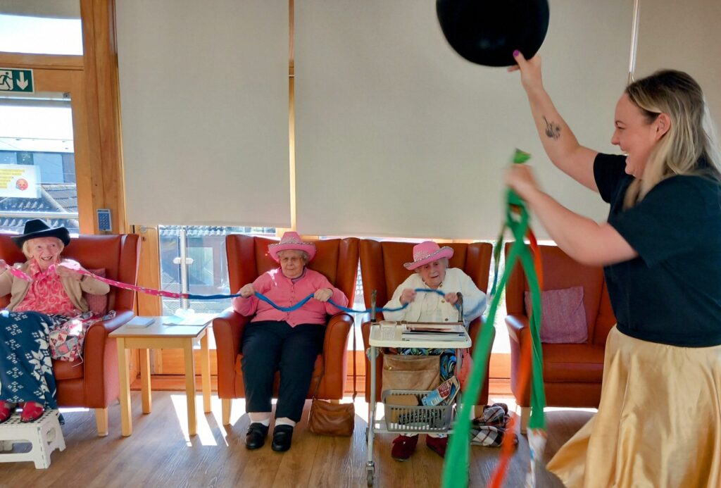 A woman waves colourful ribbons while three older women in hats sit on chairs holding the ribbons, enjoying a lively indoor Yee-Haw group activity.