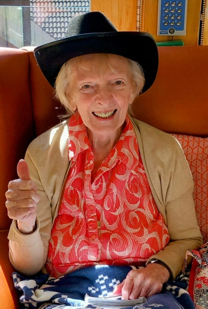 Elderly woman with fair hair wearing a black hat and patterned red shirt, sitting on an orange seat, smiling and giving a thumbs-up gesture worthy of a ‘Yee-Haw’.
