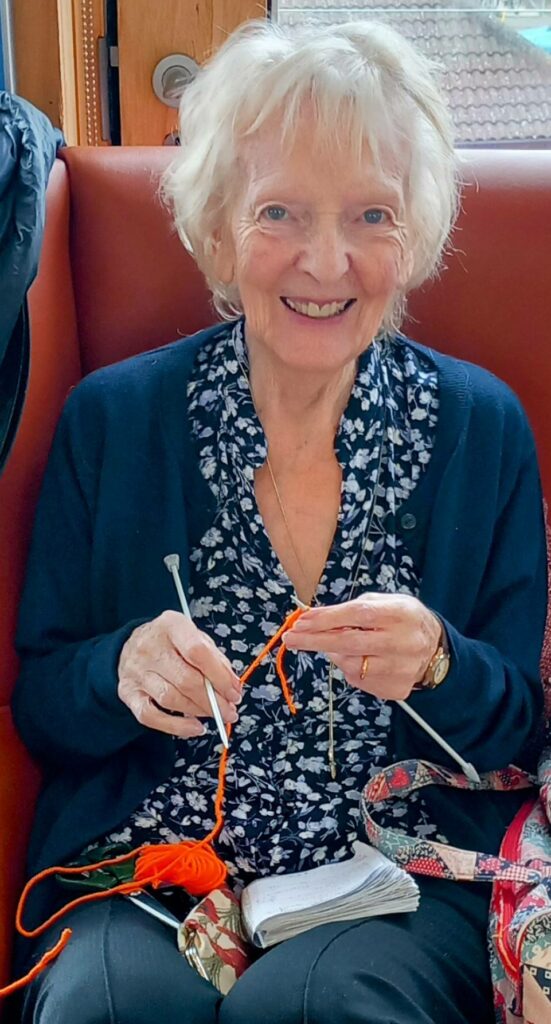 An older woman sits on a bench, smiling and knitting with orange wool and knitting needles, happily absorbed in her knitting while wearing a dark cardigan and patterned blouse.