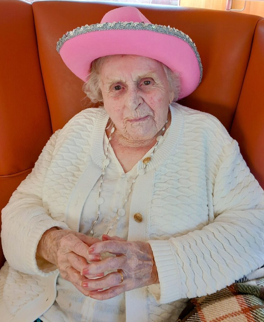Elderly woman wearing a white cardigan and a pink hat with silver trim, sitting cowboy-style on an orange chair with her hands folded.
