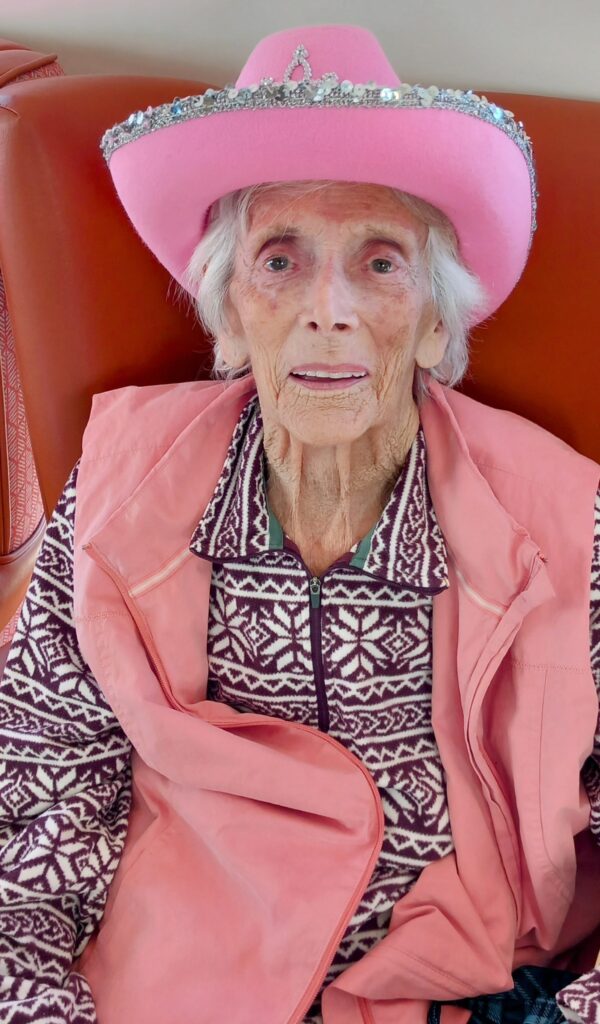 An elderly person with white hair sits on a brown chair, wearing a pink cowboy hat, patterned jumper, and pink waistcoat, expressing a Yee-Haw spirit for the camera.