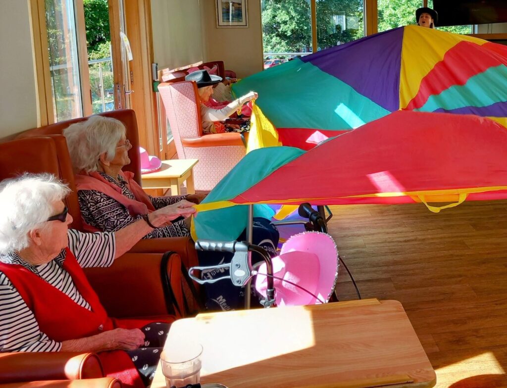 Several elderly people are seated in armchairs indoors, taking part in a lively Yee-Haw group activity with a colourful parachute, led by an individual who is partially visible in the background.