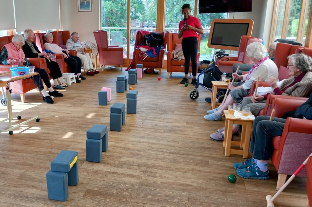 A group of elderly people sit in a semicircle in a care home while Carla stands, leading an active afternoon game using foam blocks and balls arranged on the floor.