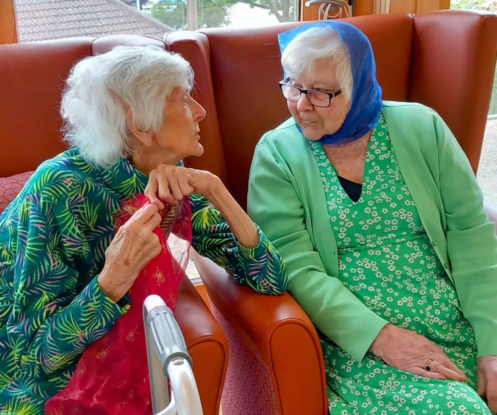 Two elderly women sit closely together on orange armchairs in a care home, engaged in conversation. One is wearing a green floral dress with a blue headscarf, while the other is dressed in a colourful top and holding a walking frame.