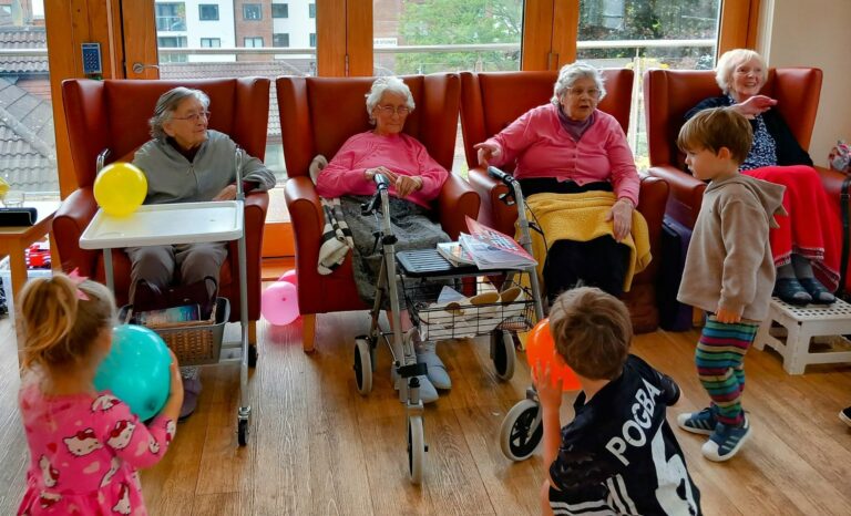 Four elderly women relax in armchairs while four young children enjoy intergenerational fun, playing with balloons in front of them in a bright, communal room at the residential care home. Rewritten in British English: Four elderly women relax in armchairs while four young children enjoy intergenerational fun, playing with balloons before them in a bright, communal lounge at the care home.